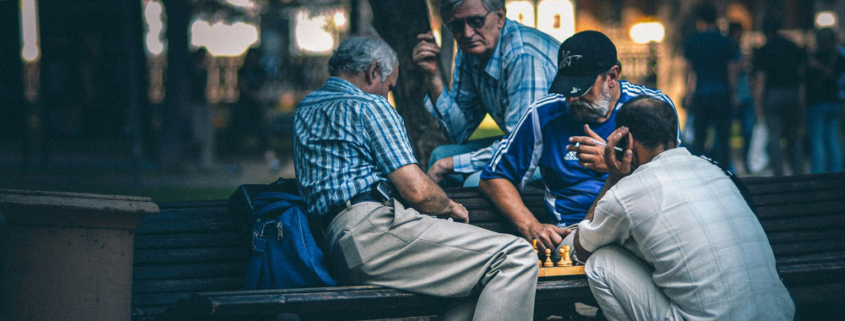 Vier ältere Männer sitzen auf einer Bank, knien davor und dahinter und spielen Schach. Zwei spielen, zwei schauen zu.